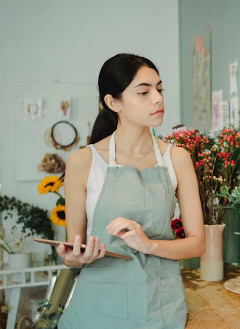 A young woman with long dark hair, wearing a light grey apron over a white sleeveless top, stands in a flower shop holding a smartphone in her right hand. She appears to be examining or organizing flo