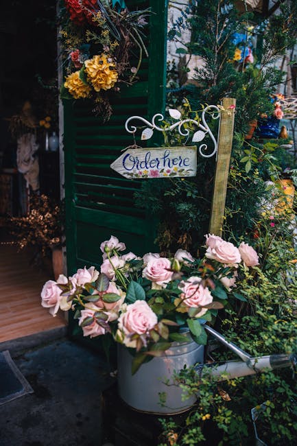 A floral display featuring a bouquet of pale pink roses with lush green leaves placed in a silver watering can, situated outdoors among various green plants and blooming flowers. In the background, a 