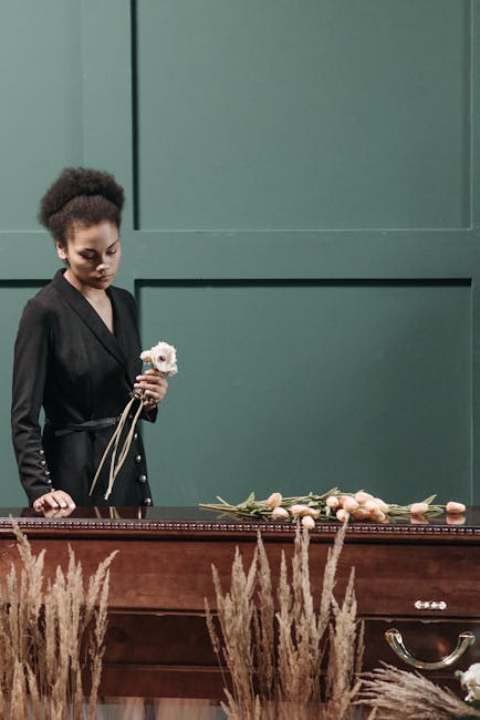 A woman with dark, curly hair styled in a puff, dressed in a black outfit, stands beside a wooden casket in a funeral setting. She holds a small white flower with pink accents, likely a rose or peony,