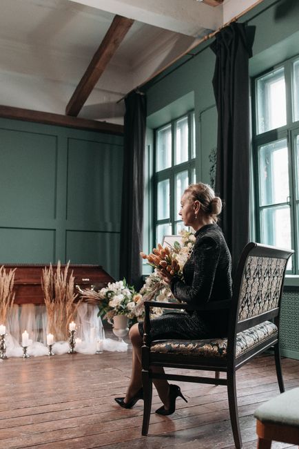 A woman seated on an elegant upholstered chair in a softly lit room with muted green walls and large windows draped in dark curtains. She is holding a bouquet of fresh flowers featuring soft pink, whi
