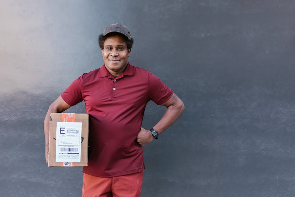 A smiling delivery person with medium brown skin and curly black hair, wearing a maroon polo shirt, matching cap, and black wristwatch, stands outdoors against a plain dark grey wall. They are holding