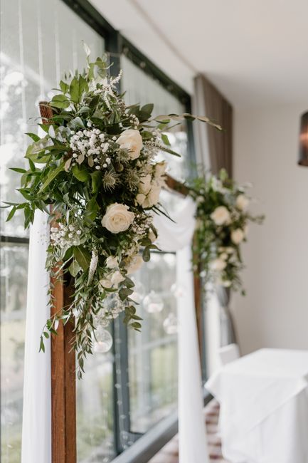 A close-up view of two elegant floral arrangements mounted on tall, dark wooden stands inside a bright room with large windows. The arrangements feature a combination of white roses, delicate baby's b