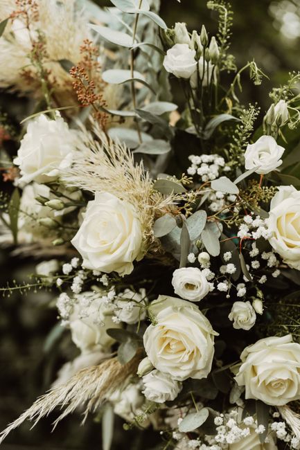 A close-up image of a floral arrangement featuring large, creamy white roses with soft, velvety petals, complemented by delicate white ranunculus and sprigs of baby's breath. The bouquet is accented w